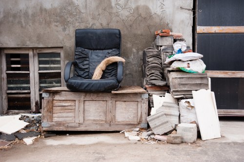 The image shows a discarded office chair with black upholstery and a cushioned seat, positioned on a wooden pallet, against a weathered concrete wall in an outdoor area. To the right of the chair, there is a pile of construction debris, including broken concrete blocks, bricks, and white plasterboard panels, some stacked atop each other and others scattered on the ground. Several folded and crumpled plastic bags are seen atop the debris, containing miscellaneous waste materials. Behind the debris, part of a black metal structure or gate is visible, suggesting an industrial or backstreet setting in Peckham. The lighting appears natural and subdued, emphasizing the rough texture of the wall and the materials' worn appearances, typical of waste awaiting collection or removal by a local rubbish clearance service such as Office Clearance Peckham.

This scene exemplifies typical rubbish found in an urban process of clearing or disposing of unwanted office or construction materials in the Peckham area of south London, aligning with services aimed at local rubbish removal and clearance solutions.