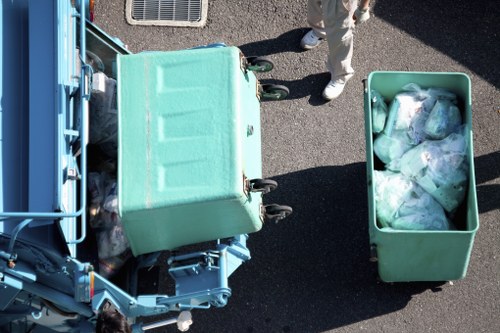 An overhead view of a teal-colored waste collection bin positioned on a paved street or parking area, partially open with transparent plastic bags filled with assorted rubbish visible inside. The bin is situated next to a larger teal-colored waste container or recycling bin, which is also slightly open, revealing more rubbish inside. To the right of the bins, a person dressed in light-colored clothing and white shoes is standing nearby, with only their legs visible in the image. The surrounding environment appears to be an outdoor urban or commercial area, with the street surface clearly visible beneath the bins, and shadows suggesting it is a sunny day. The scene reflects a typical waste management or rubbish removal scenario, consistent with services offered by Office Clearance Peckham in the Peckham area.