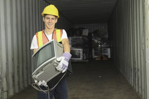 A young man wearing a yellow safety helmet, high-visibility orange and yellow vest, white T-shirt, and gloves is standing inside a large shipping container with metal walls. He is holding an old, bulky CRT television with a grey plastic casing, a curved screen, and visible cables hanging from the back. The container's interior appears industrial, with dark shadows at the far end and some piled rubbish or discarded appliances faintly visible in the background. The scene is set outdoors or in an industrial area, reflecting a rubbish removal or clearance activity typical of services offered by Office Clearance Peckham, which operates in the Peckham area and nearby postcode regions. The overall scene emphasizes waste collection and appliance disposal, relevant to local rubbish clearance services in London, particularly within the South London district.