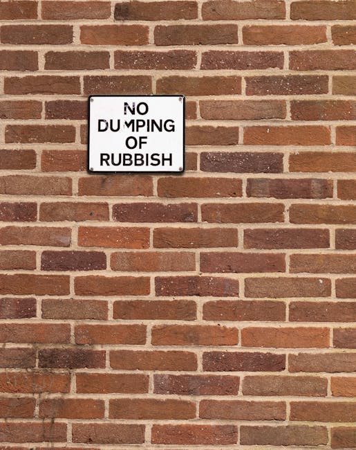 A white rectangular metal sign mounted on a red brick wall displaying the message 'NO DUMPING OF RUBBISH' in black capital letters with some minor fading and chipping around the edges. The brick wall
