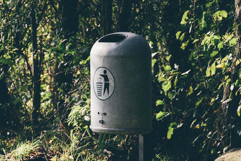 A metallic rubbish collection box, positioned on a grassy verge adjacent to a paved pathway in a park setting. The box has a rectangular shape with a slightly weathered, light grey surface, showing so